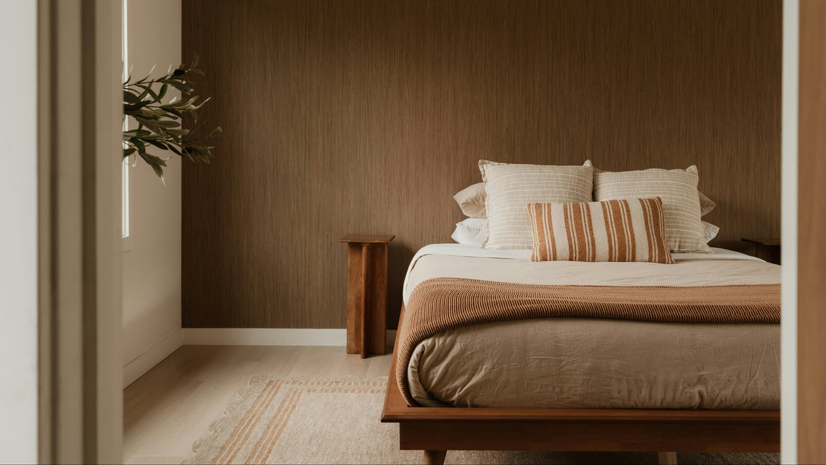 A modern bedroom interior with a low wooden bed frame, neutral beige bedding, striped accent pillows, and a dark brown wood-paneled wall. The photo is framed by a doorway, with a small plant visible on the left.
